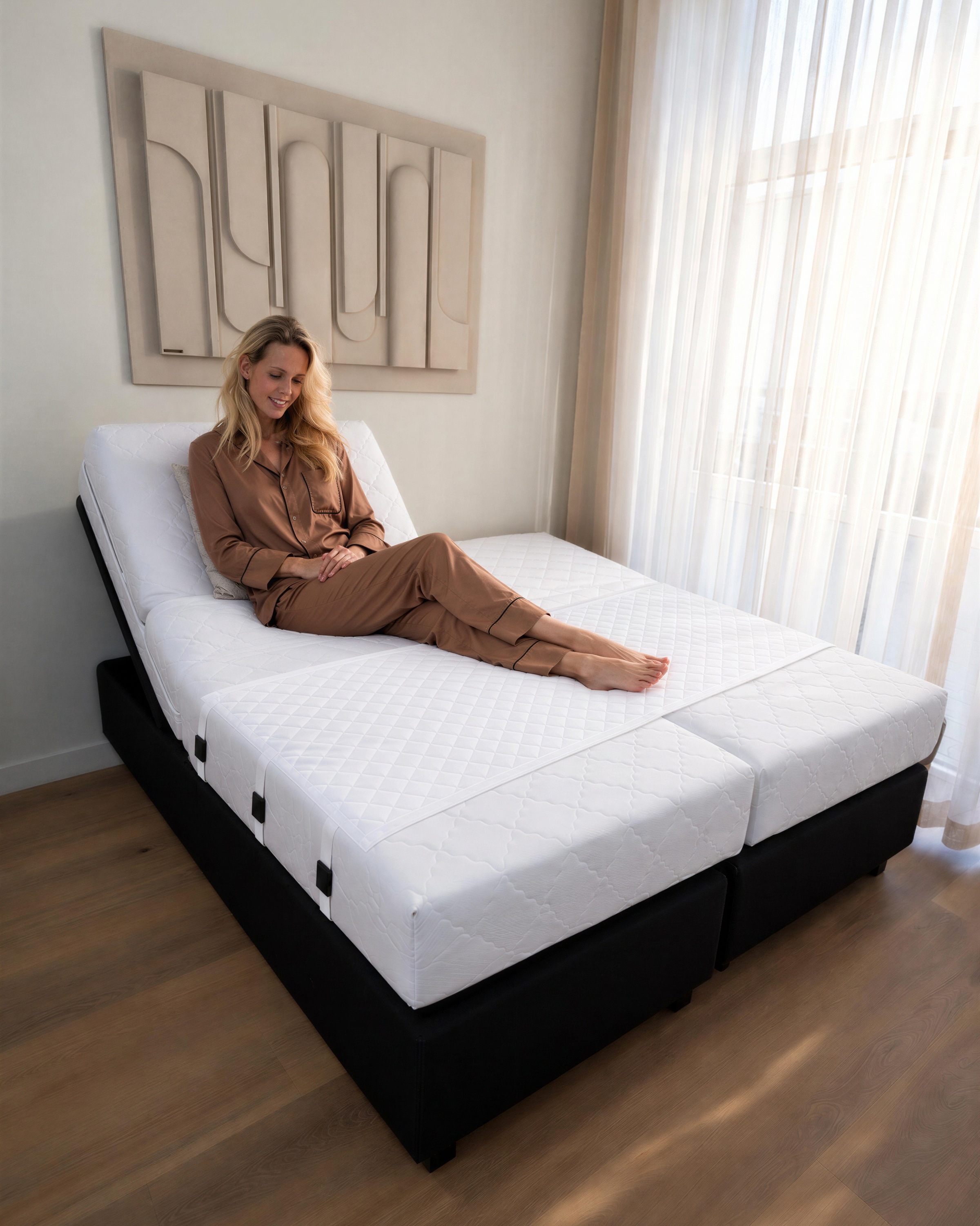 Une femme assise sur un lit électrique à deux matelas, la tête relevée, les trois sangles du Bedbinder visibles sur le côté